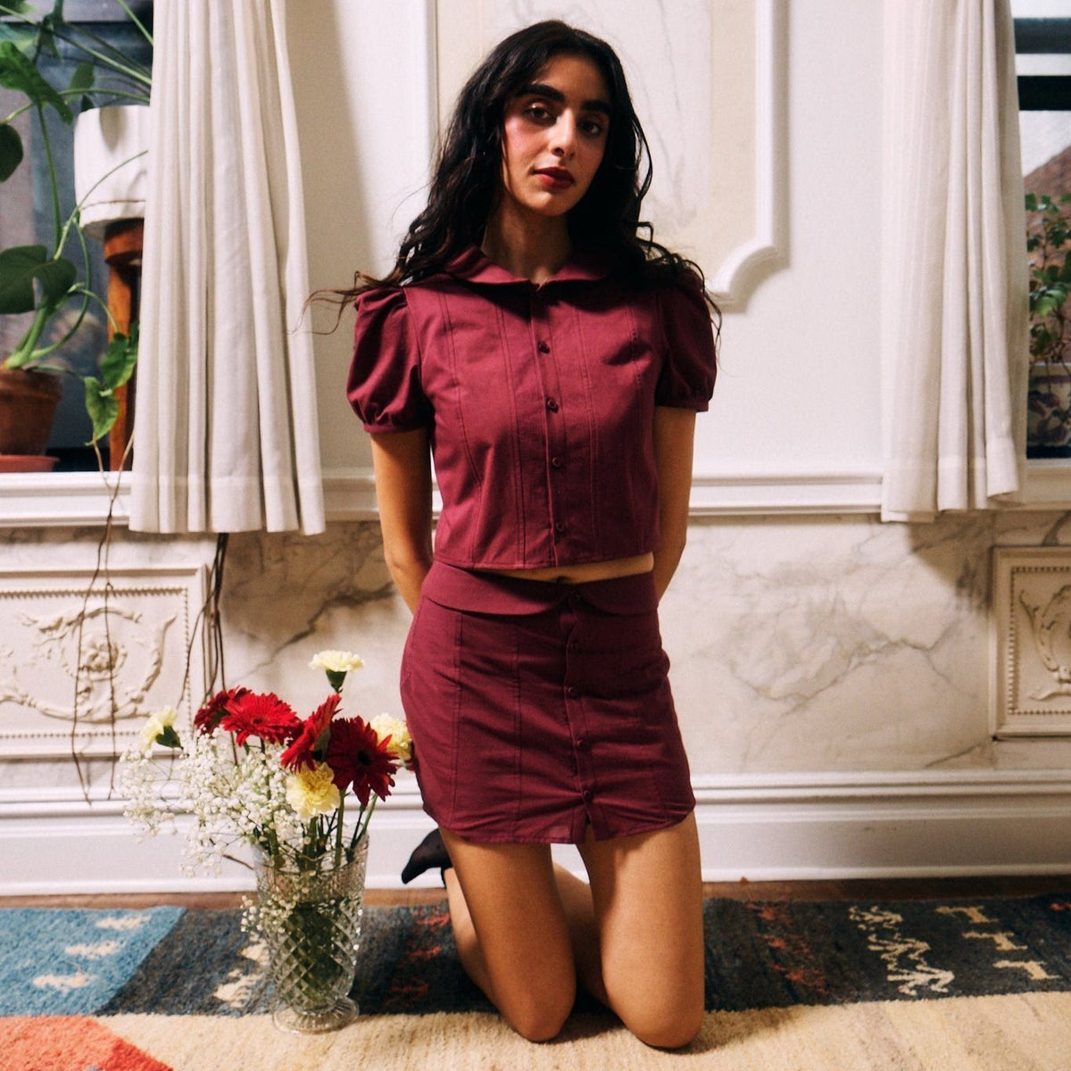 Enketta Skirt and Blouse in pomegranate, model kneeling on a rug beside a vase of flowers