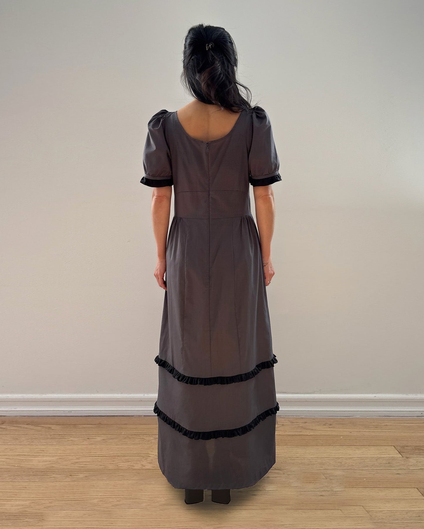 Woman wearing a long, dark dress with short sleeves and black trim against a plain wall.