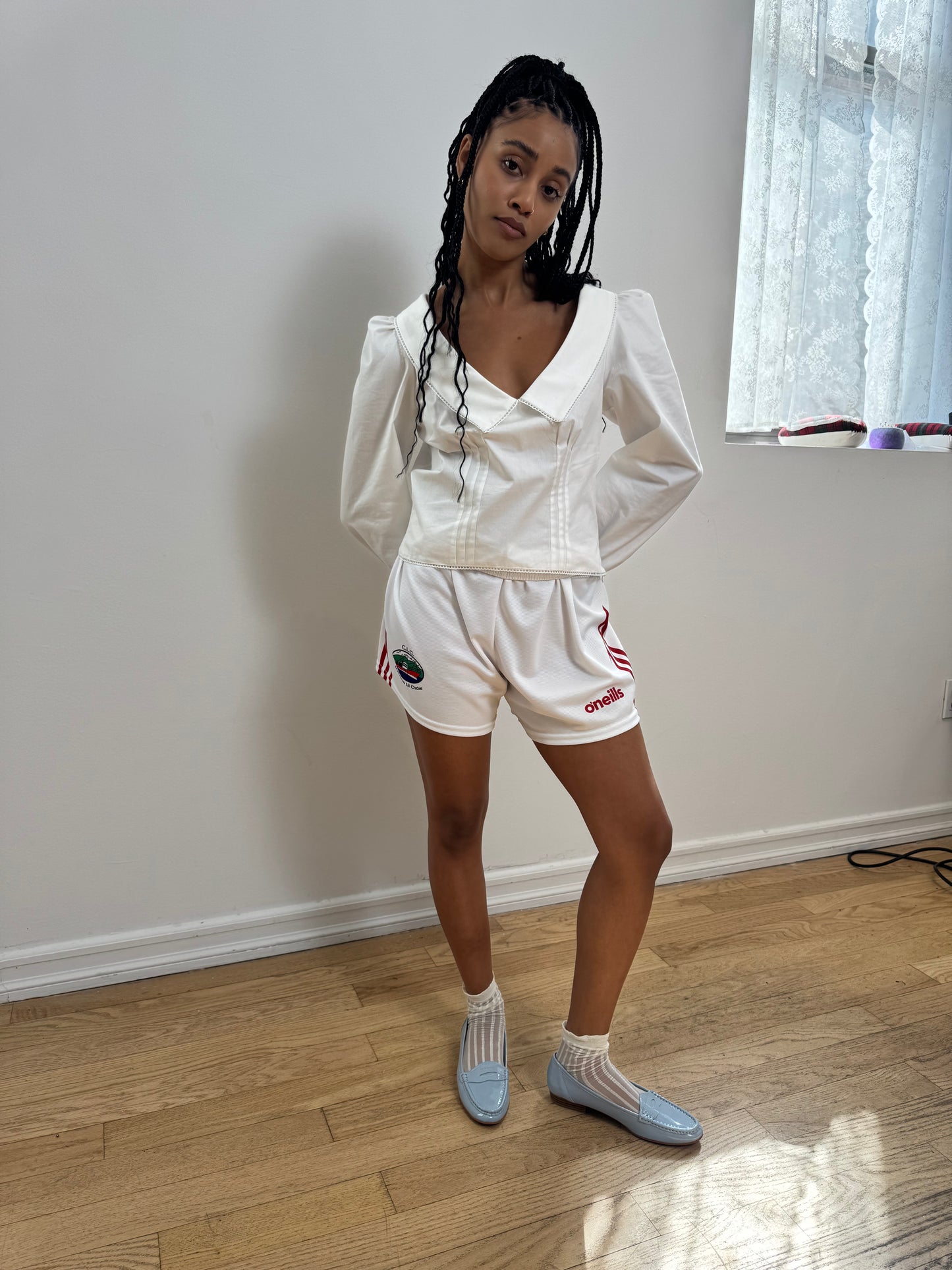 A woman stands in a room with light wood floors and a white wall, wearing a white long-sleeve top and white gym shorts with small red and green logo details, paired with white socks and light blue slip-on shoes.