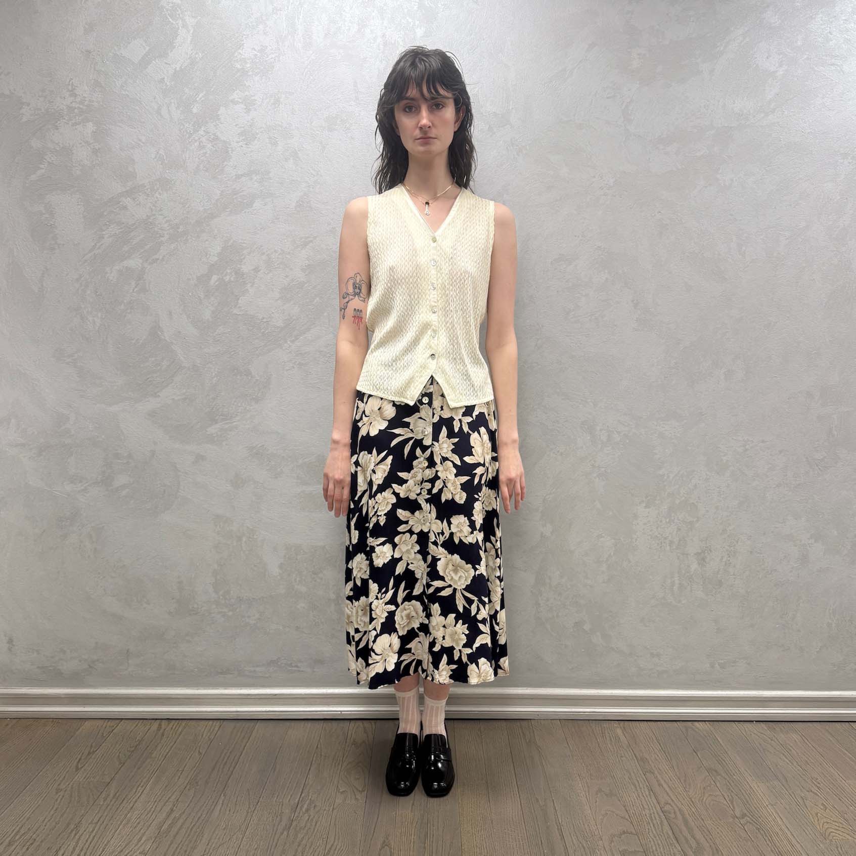 Person wearing a white sleeveless top and floral skirt against a plain wall.