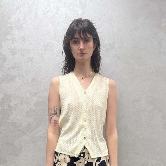 Person wearing a white sleeveless top with buttons down the front and floral skirt against a plain wall.
