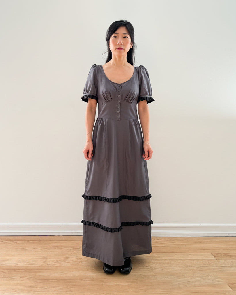Woman wearing a long gray dress with black trim against a plain wall.