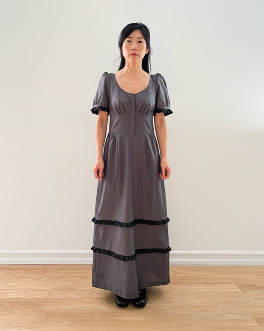 Woman wearing a long gray dress with black trim against a plain wall.